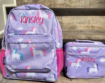 May include: A purple backpack and matching lunch bag set featuring a unicorn pattern. The backpack and lunch bag have the name "Kinsley" embroidered in pink. The set includes pink mesh side pockets.