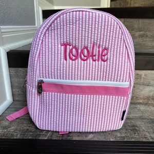 May include: Pink and white striped backpack with the name "Toolie" embroidered in pink. The backpack has a zippered front pocket with a white zipper and a pink strap. The bag is sitting on a wooden surface.