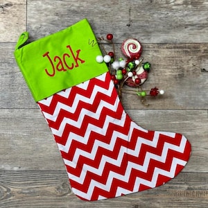 May include: A red and white chevron Christmas stocking with a green cuff. The cuff is embroidered with the name "Jack".