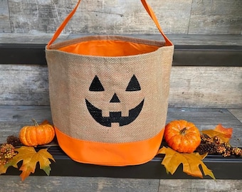 May include: Orange and brown burlap trick-or-treat bag with a black jack-o'-lantern face. The bag is sitting on a wooden step with two small orange pumpkins and fall foliage.