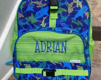 May include: A green and blue backpack with a shark pattern. The backpack has the name "ADRIAN" embroidered on the front. It features a top handle, adjustable straps, and a buckle closure. The backpack is designed for carrying school supplies.