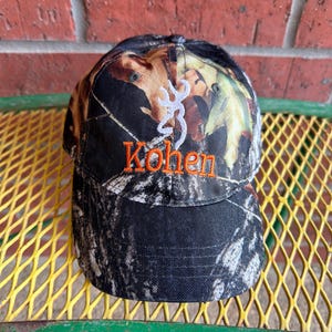 May include: A camouflage baseball cap featuring a white embroidered deer antler logo above the orange embroidered name Kohen. The cap has a structured crown and a curved brim, displaying a woodland pattern with shades of black, brown, and green.