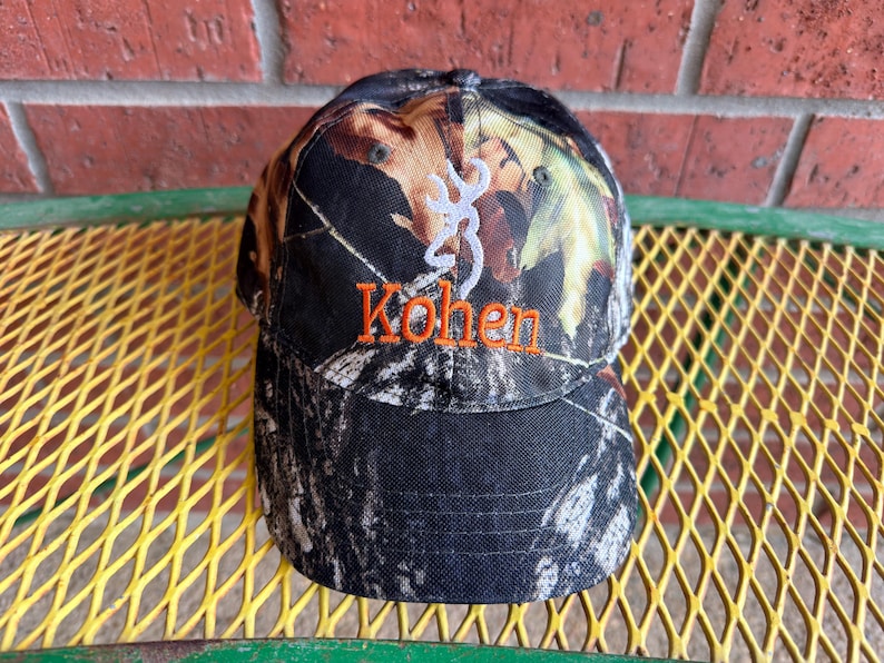 May include: A camouflage baseball cap featuring a white embroidered deer antler logo above the orange embroidered name Kohen. The cap has a structured crown and a curved brim, displaying a woodland pattern with shades of black, brown, and green.