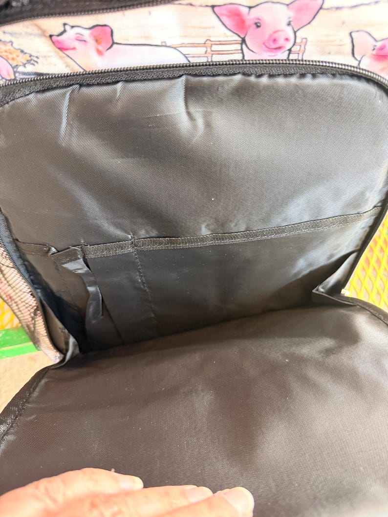 Puede incluir: Mochila negra con un estampado de cerdos. El interior es negro con m&uacute;ltiples compartimentos. El exterior presenta cerdos rosas con un dise&ntilde;o de valla de madera. La mochila tiene una cremallera negra y un bolsillo lateral de malla.