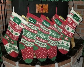 May include: Five knitted Christmas stockings featuring a red, green, and white fair isle pattern with snowflakes, trees, and gift motifs. Each stocking has a green cuff embroidered with the names Mom, Ethan, Henrik, Gallen, and Dad respectively.