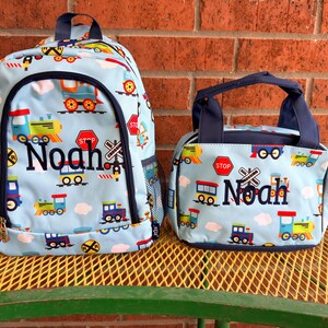May include: A light blue backpack and matching lunch bag set featuring a colorful train, railroad crossing sign, and stop sign pattern. Both items are personalized with the name Noah embroidered in dark blue thread on the front panels.