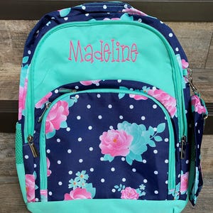 May include: A blue and green backpack with white polka dots and floral designs. The backpack has a green zippered pocket on the front with the name "Madeline" embroidered in pink.