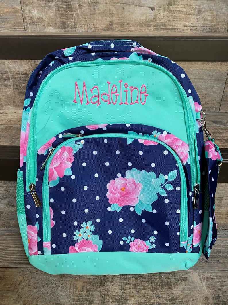 May include: A blue and green backpack with white polka dots and floral designs. The backpack has a green zippered pocket on the front with the name "Madeline" embroidered in pink.