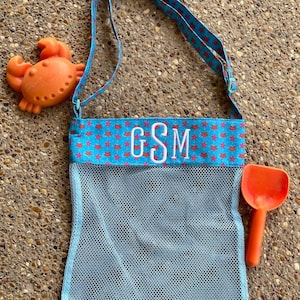 May include: A blue mesh beach bag with a red and blue crab print strap. The bag has a white monogram "GSM" on the front. A red plastic shovel and a red plastic crab toy are next to the bag.