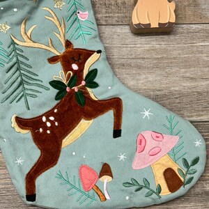 May include: A sage green Christmas stocking featuring an embroidered brown deer with antlers, a green wreath around its neck, and pink and brown mushrooms. The stocking is decorated with gold glitter stars, white embroidered snowflakes, and green pine trees.