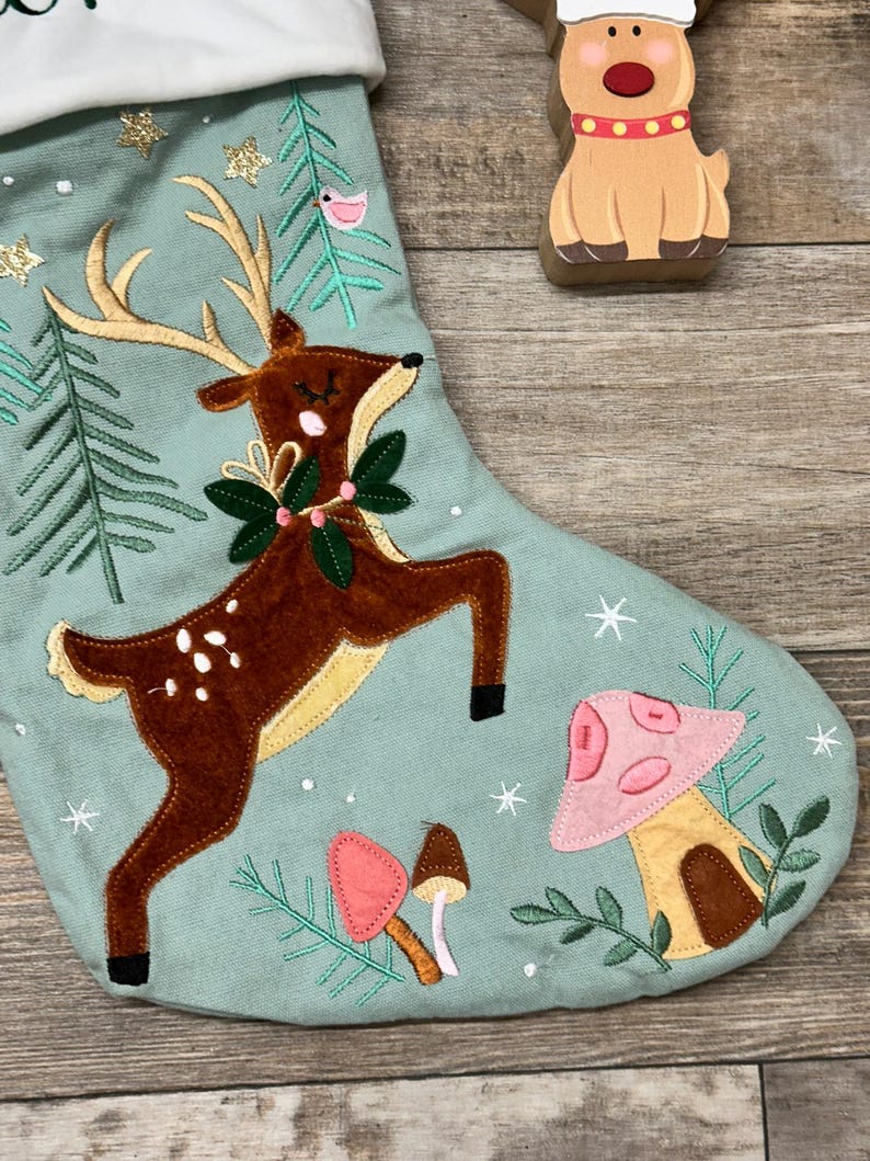 May include: A sage green Christmas stocking featuring an embroidered brown deer with antlers, a green wreath around its neck, and pink and brown mushrooms. The stocking is decorated with gold glitter stars, white embroidered snowflakes, and green pine trees.