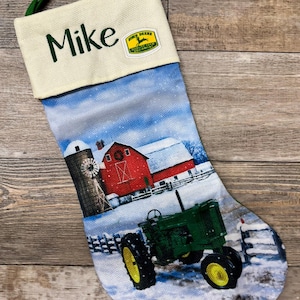 May include: A personalized Christmas stocking with a winter farm scene featuring a red barn, a windmill, and a green John Deere tractor. The stocking is white with a cream-colored cuff that has the name "Mike" embroidered in green and a John Deere logo patch.