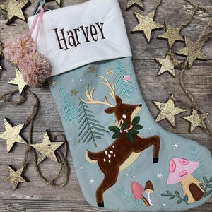 May include: A light blue Christmas stocking with a white cuff embroidered with the name Harvey. The stocking features a brown deer with antlers, a green wreath, pink and brown mushrooms, and pine tree embroidery, accented by two pink pom-poms.