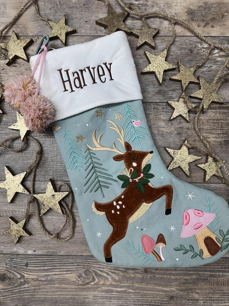 May include: A light blue Christmas stocking with a white cuff embroidered with the name Harvey. The stocking features a brown deer with antlers, a green wreath, pink and brown mushrooms, and pine tree embroidery, accented by two pink pom-poms.