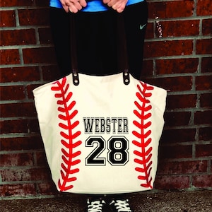 K&ouml;nnte beinhalten: Eine wei&szlig;e Canvas-Tragetasche mit einem roten Baseball-Design und dem Namen "Webster" und der Nummer "28" in Schwarz gedruckt.