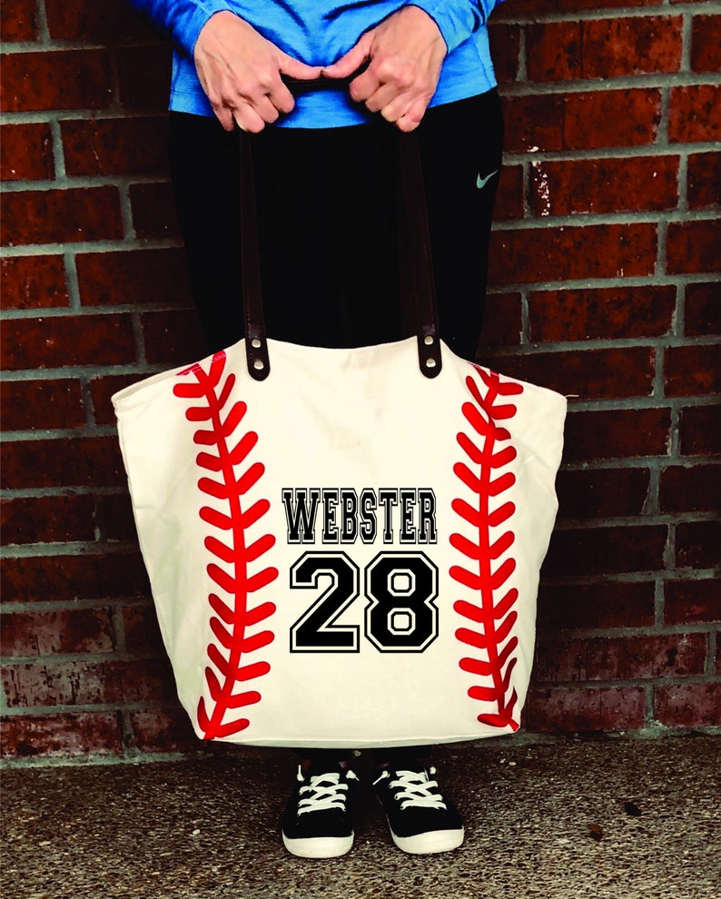 K&ouml;nnte beinhalten: Eine wei&szlig;e Canvas-Tragetasche mit einem roten Baseball-Design und dem Namen "Webster" und der Nummer "28" in Schwarz gedruckt.