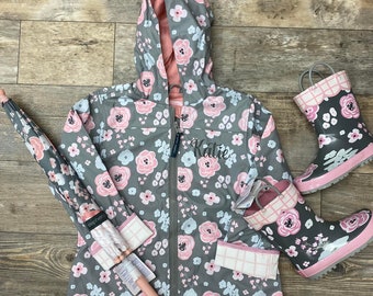 May include: A gray hooded raincoat with a pink floral pattern and the name "Katie" embroidered on the front. The raincoat is paired with a pink umbrella and pink rain boots with a gray floral pattern.