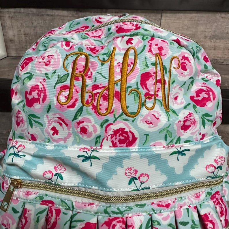 May include: A floral-patterned backpack with a light blue background and pink roses. The backpack features gold-colored embroidered initials "RHN" and a gold zipper. The bottom of the backpack has a pleated design.