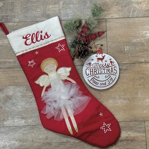 May include: Red Christmas stocking with a white angel and gold glitter accents. The stocking is personalized with the name "Ellis" and has a white ornament with the text "Merry Christmas Peace and Joy".
