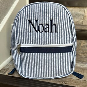 May include: A blue and white striped backpack with the name "Noah" embroidered on the front. The backpack has a zippered pocket, navy blue accents, and adjustable straps. The backpack is made of a textured fabric.