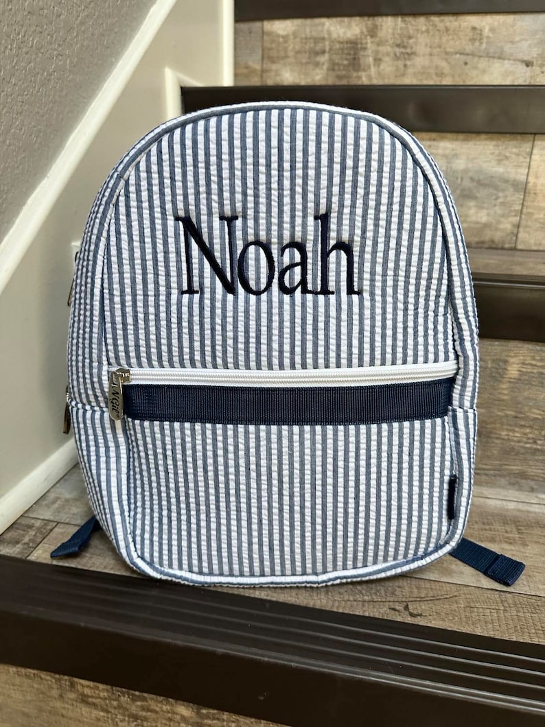 May include: A blue and white striped backpack with the name "Noah" embroidered on the front. The backpack has a zippered pocket, navy blue accents, and adjustable straps. The backpack is made of a textured fabric.