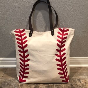 K&ouml;nnte beinhalten: Eine wei&szlig;e Canvas-Tragetasche mit roten Baseballn&auml;hten. Die Tasche hat braune Ledergriffe und Metall-Akzente.