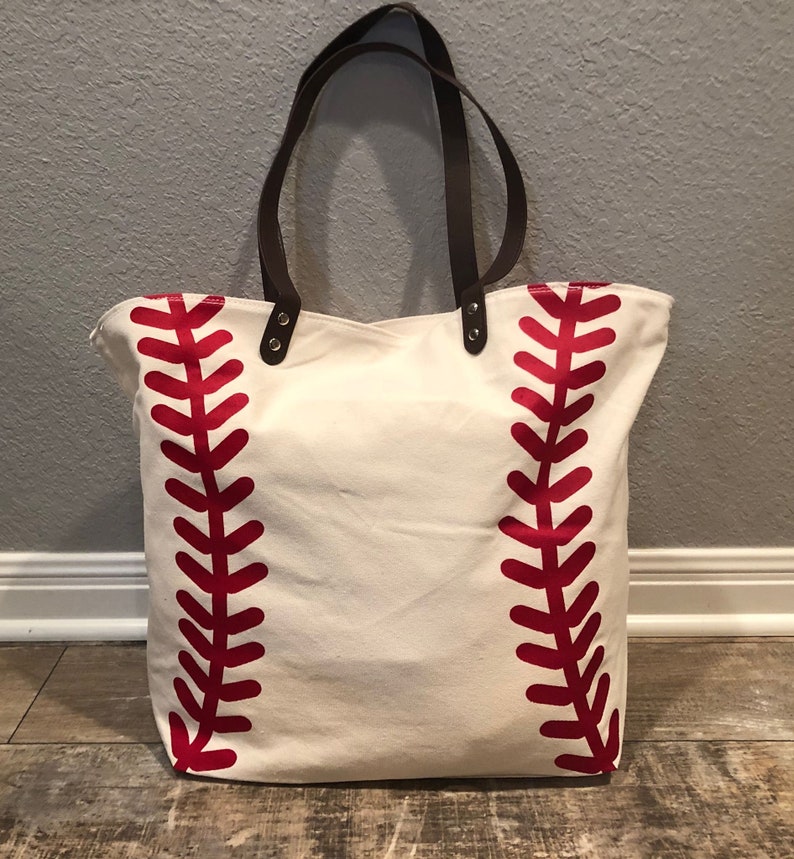 K&ouml;nnte beinhalten: Eine wei&szlig;e Canvas-Tragetasche mit roten Baseballn&auml;hten. Die Tasche hat braune Ledergriffe und Metall-Akzente.