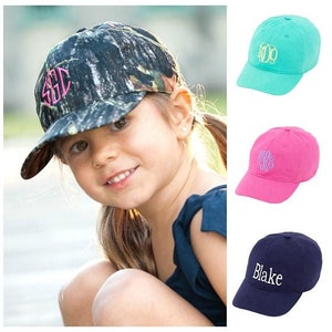 May include: Three different styles of baseball caps with embroidered initials. The caps are in camouflage, teal and pink.