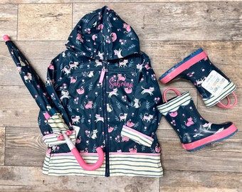 May include: A blue raincoat with a pink zipper and a white and pink striped trim. The raincoat has a hood and is decorated with a pattern of pink cats and flowers. The raincoat is personalized with the name "Sabrina". The image also includes a pink and blue umbrella and a pair of pink and blue rain boots with a cat pattern.