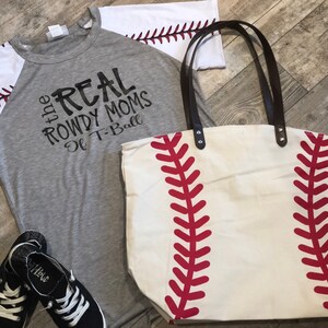 K&ouml;nnte beinhalten: Ein graues T-Shirt mit wei&szlig;en &Auml;rmeln und dem Aufdruck "the REAL ROWDY MOMS of T-Ball" auf der Vorderseite. Neben dem Shirt befindet sich eine wei&szlig;e Stofftasche mit roten Baseballn&auml;hten.