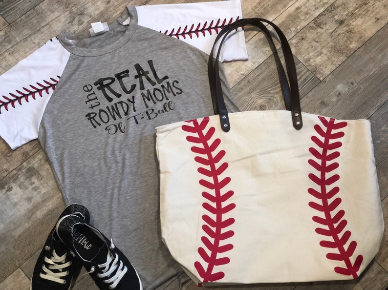 K&ouml;nnte beinhalten: Ein graues T-Shirt mit wei&szlig;en &Auml;rmeln und dem Aufdruck "the REAL ROWDY MOMS of T-Ball" auf der Vorderseite. Neben dem Shirt befindet sich eine wei&szlig;e Stofftasche mit roten Baseballn&auml;hten.