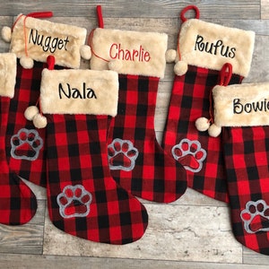 May include: Six red and black buffalo plaid Christmas stockings with white faux fur trim and a paw print design. The stockings are personalized with the names Lulu, Nugget, Nala, Charlie, Rufus, and Bowie.