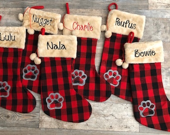May include: Six red and black buffalo plaid Christmas stockings with white faux fur trim and a paw print design. The stockings are personalized with the names Lulu, Nugget, Nala, Charlie, Rufus, and Bowie.