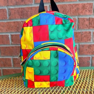May include: A colorful backpack with a pattern resembling toy building blocks. The backpack features a main compartment and a front pocket, both with black zippers. The design incorporates red, yellow, blue, and green blocks, with a black handle and straps.