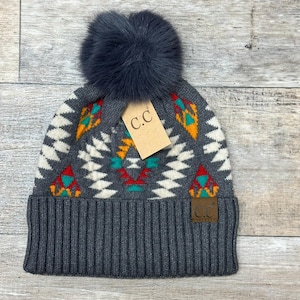 May include: Gray knit beanie with a colorful geometric pattern and a fluffy gray pom pom. The beanie has a brown leather patch with the letters "C.C." on it.