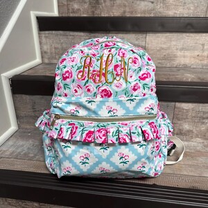 May include: A floral-patterned backpack with pink roses and a light blue background. The backpack features a gold zipper, ruffled trim, and embroidered initials. The bag has a top handle and adjustable straps.