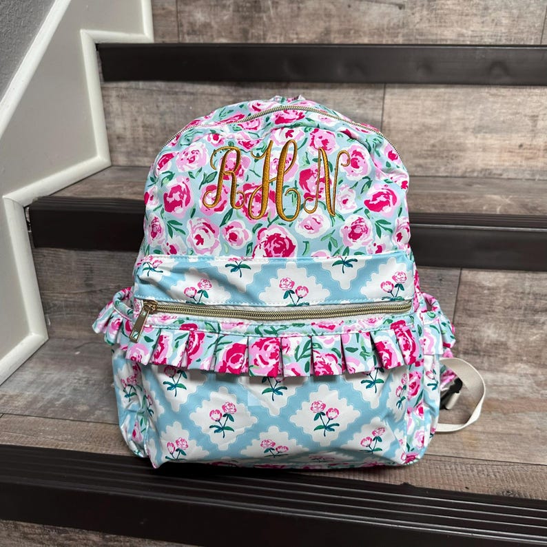 May include: A floral-patterned backpack with pink roses and a light blue background. The backpack features a gold zipper, ruffled trim, and embroidered initials. The bag has a top handle and adjustable straps.