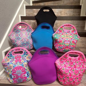 May include: A collection of colorful lunch bags in various designs and colors. The bags include solid colors like black, blue, and purple, and patterned bags with floral and geometric designs. Each bag has a handle.