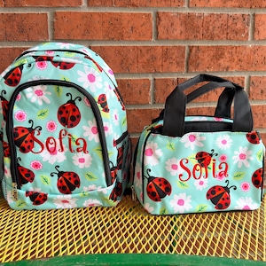 May include: A light blue backpack and a matching lunch bag, both featuring a repeating pattern of red ladybugs and white flowers. Each item is personalized with the name Sofia in red cursive script. The backpack has black mesh side pockets.