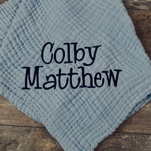May include: A light blue muslin blanket with a textured, crinkled fabric surface. The name Colby Matthew is embroidered in dark navy blue thread across the center of the blanket in a bold, decorative script font with clear, legible lettering.