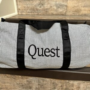 May include: A black and white striped duffel bag with black straps and the word "Quest" embroidered in black on the front.