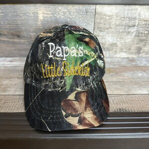 May include: Camouflage baseball cap with white embroidery that says "Papa's Little Sidekick" in yellow.