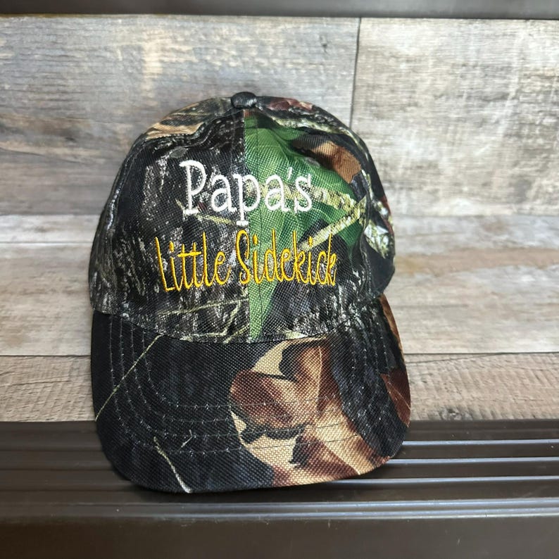May include: Camouflage baseball cap with white embroidery that says "Papa's Little Sidekick" in yellow.
