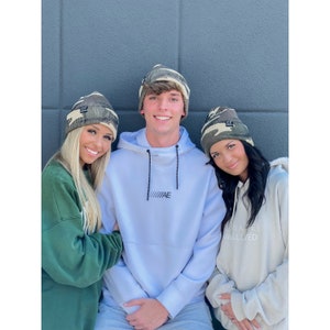 May include: Three people wearing camouflage beanies with black patches. The beanie on the left has the letters "CC" on the patch. The beanie in the middle has the letters "AE" on the patch. The beanie on the right has the letters "CC" on the patch. The person in the middle is wearing a light blue hoodie with a black zipper. The person on the right is wearing a cream colored hoodie with the text "THE LIFE WELL LIVED" on it.