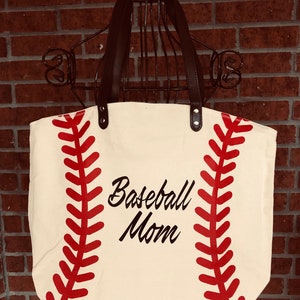 K&ouml;nnte beinhalten: Eine wei&szlig;e Canvas-Tragetasche mit schwarzen Ledergriffen. Die Tasche hat ein rotes Baseball-Nahtdesign und den Text "Baseball Mom" in Schwarz.
