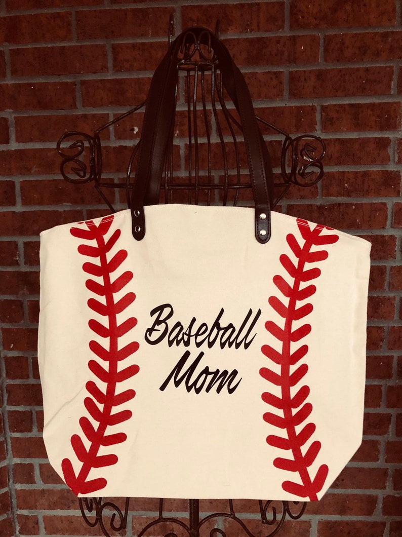 K&ouml;nnte beinhalten: Eine wei&szlig;e Canvas-Tragetasche mit schwarzen Ledergriffen. Die Tasche hat ein rotes Baseball-Nahtdesign und den Text "Baseball Mom" in Schwarz.
