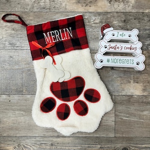 May include: Red and black buffalo plaid Christmas stocking with a white faux fur cuff and a large paw print design. The stocking is personalized with the name "MERLIN" and has a bone-shaped ornament attached. A white wooden sign with three bone-shaped tiers reads "I ate Santa's cookies #noregrets".