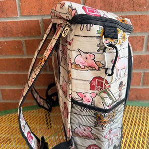 Puede incluir: Una mochila blanca y beige con un estampado de animales de granja con cerdos rosas y graneros rojos. La mochila tiene correas negras, una cremallera negra y un bolsillo lateral de malla negra. La marca "NGE" es visible.