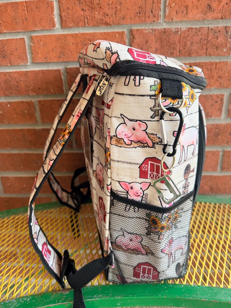 Puede incluir: Una mochila blanca y beige con un estampado de animales de granja con cerdos rosas y graneros rojos. La mochila tiene correas negras, una cremallera negra y un bolsillo lateral de malla negra. La marca "NGE" es visible.