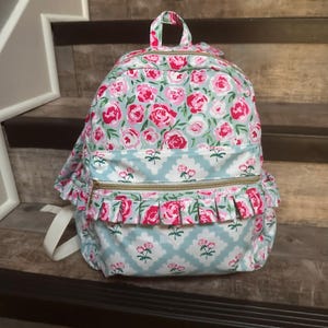 May include: A floral backpack with a light blue background and pink roses. The backpack has a top handle, two zippered compartments, and white shoulder straps. Ruffled trim and gold zippers add detail.
